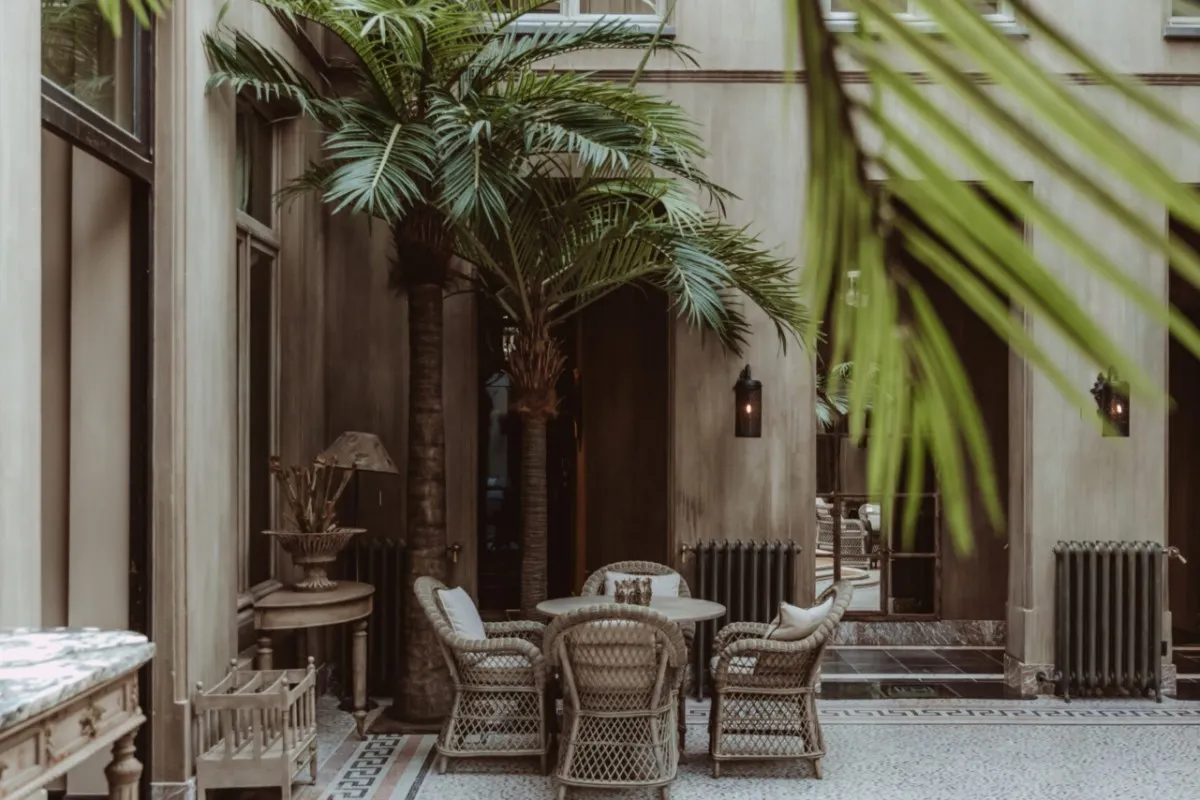 Courtyard of Cobergher Hotel with stone façade, palm trees, wicker chairs and table under arched walls.