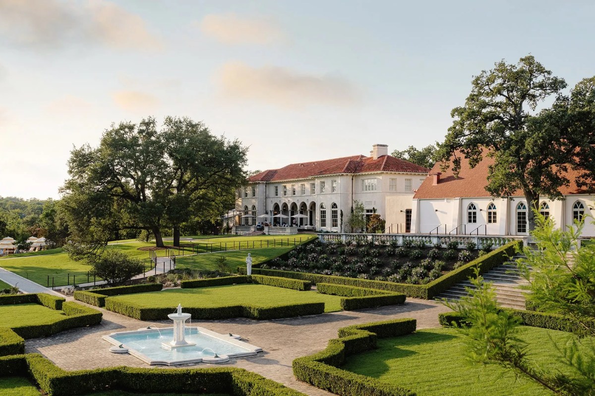 Elegant white mansion exterior with red tile roof, manicured hedge gardens and a fountain in foreground at golden hour