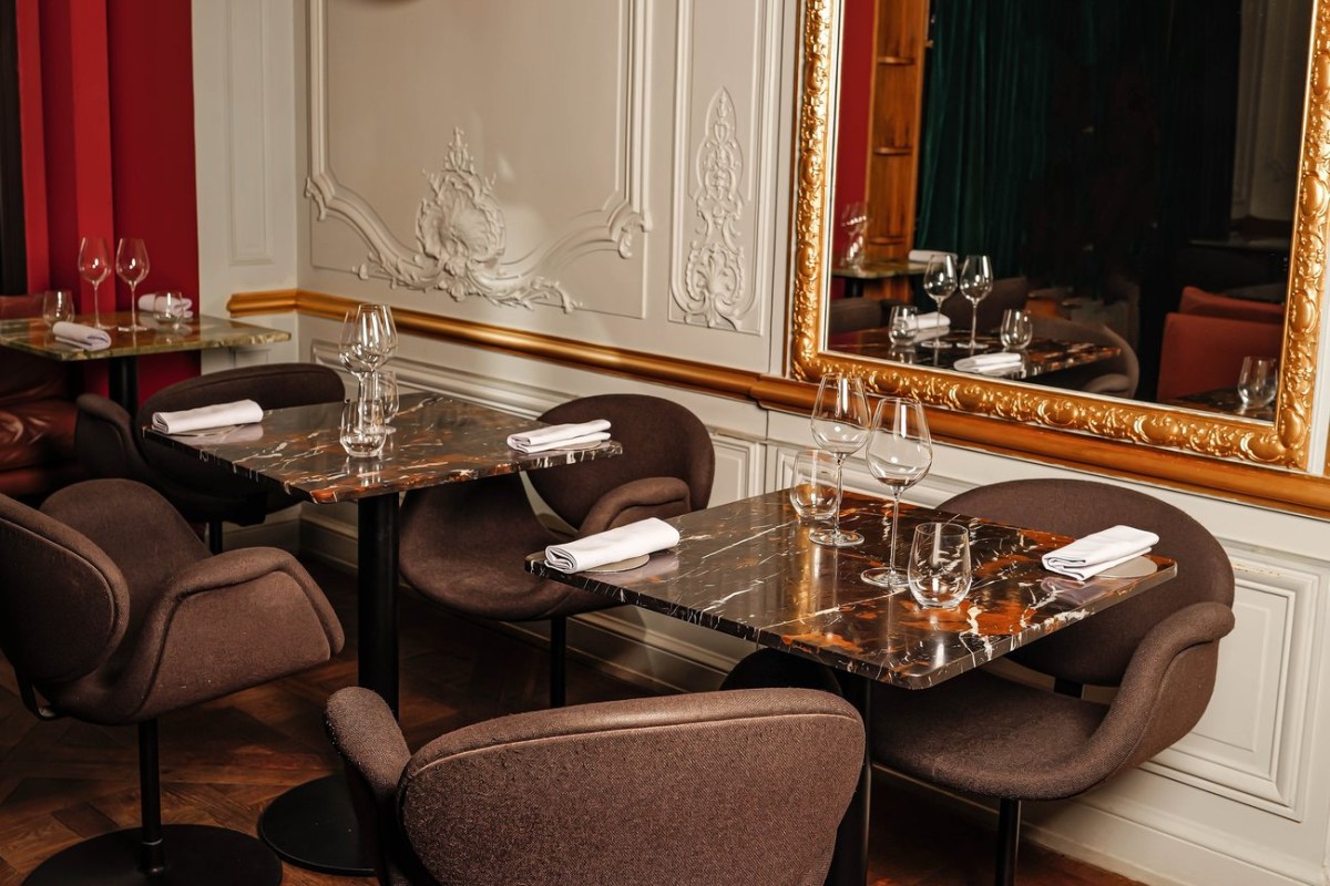 Elegant dining room with marble-top tables, ornate molding, and a large gilt mirror reflecting red velvet drapes.