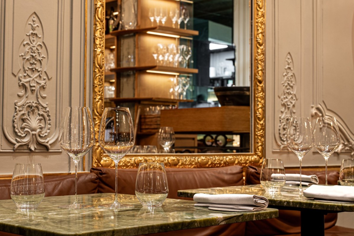 Restaurant interior with marble tables set with wine glasses and napkins, gilt-framed mirror reflecting a shelf of glassware.