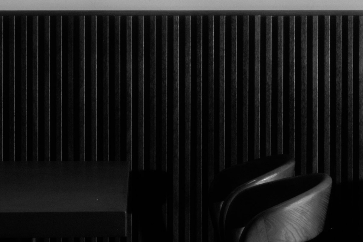 Black and white detail of dark wood slat paneling with a wooden chair and table edge in a dim dining room