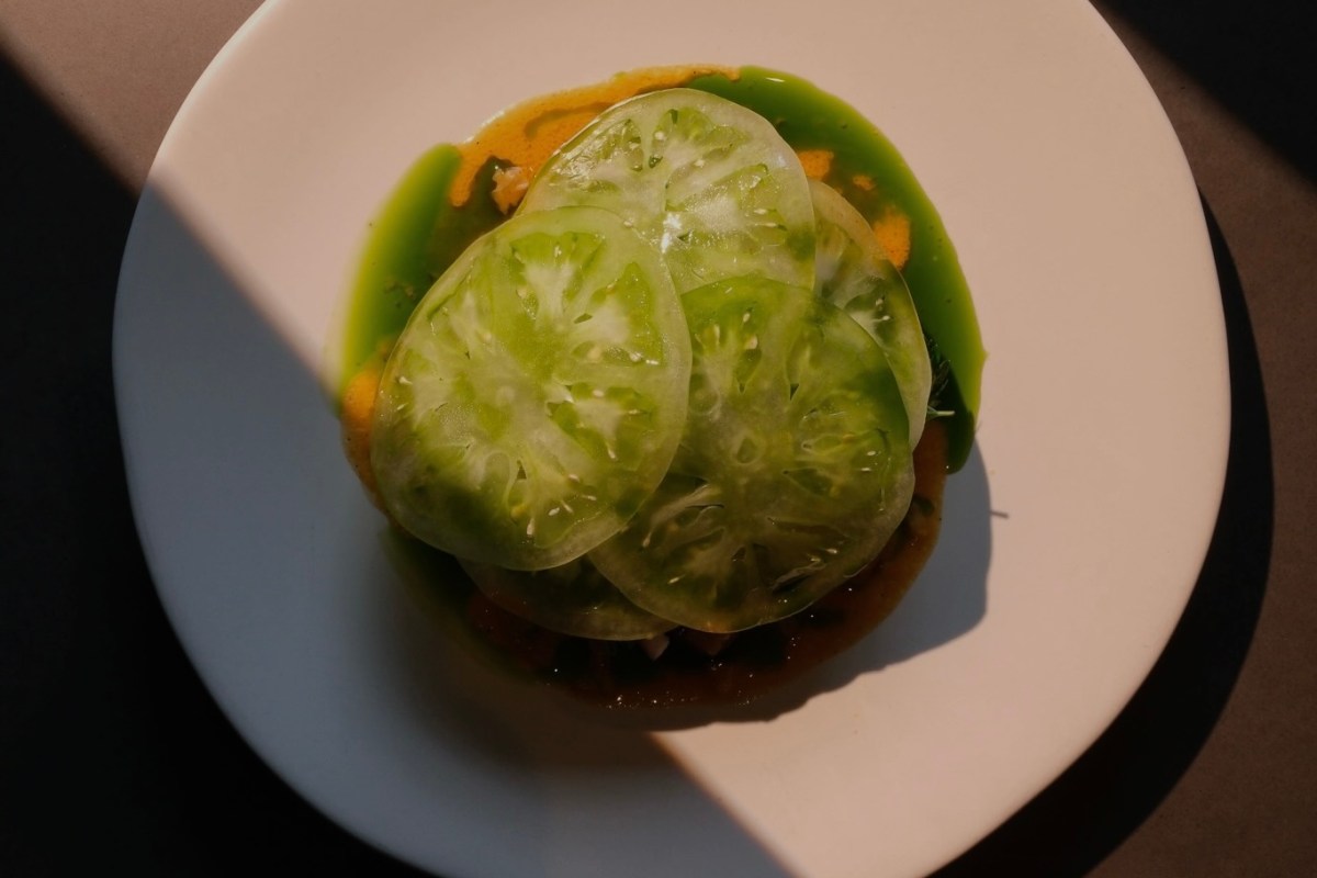 Overhead shot of sliced green tomatoes on a white plate with colorful sauces, sunlight casting shadow