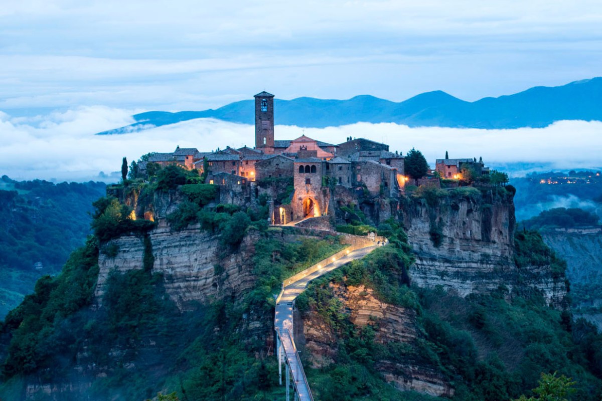 Aerial twilight view of Civita di Bagnoregio's hilltop town on a cliff, connected by a footbridge, with mountains and fog behind