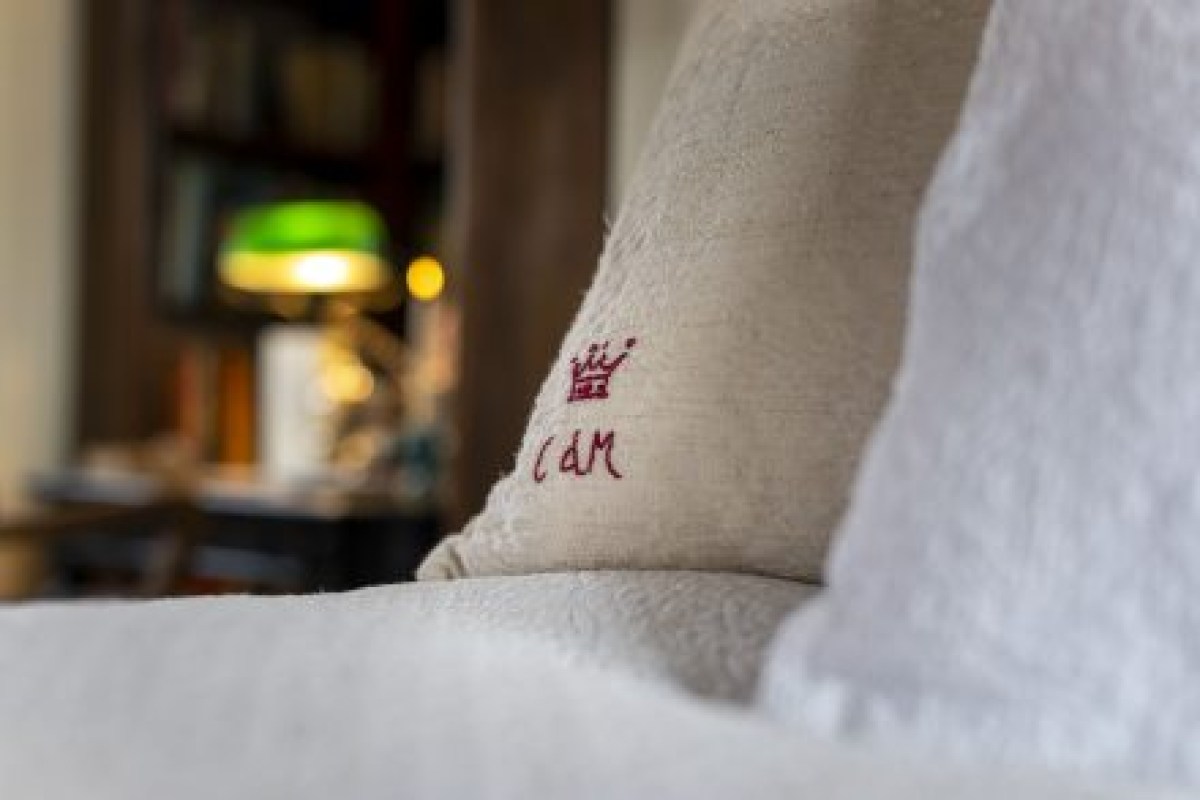 Close-up of embroidered pillow with crown logo and 'CdM' monogram, cozy interior with bookshelf and lamp in background