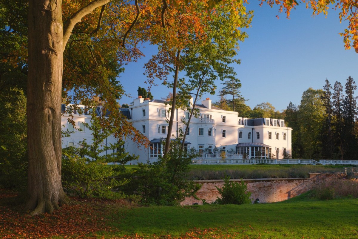 White mansion exterior of Coworth Park surrounded by autumn trees and lawn under blue sky