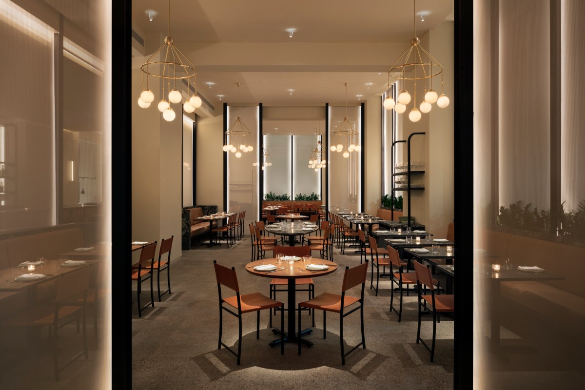 Stylish restaurant dining room viewed through glass doors, with round tables, leather chairs, and hanging globe lights.