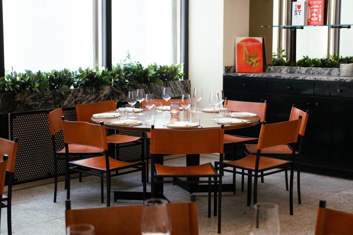 Round dining table set with wine glasses and plates, surrounded by leather chairs near large windows with greenery.