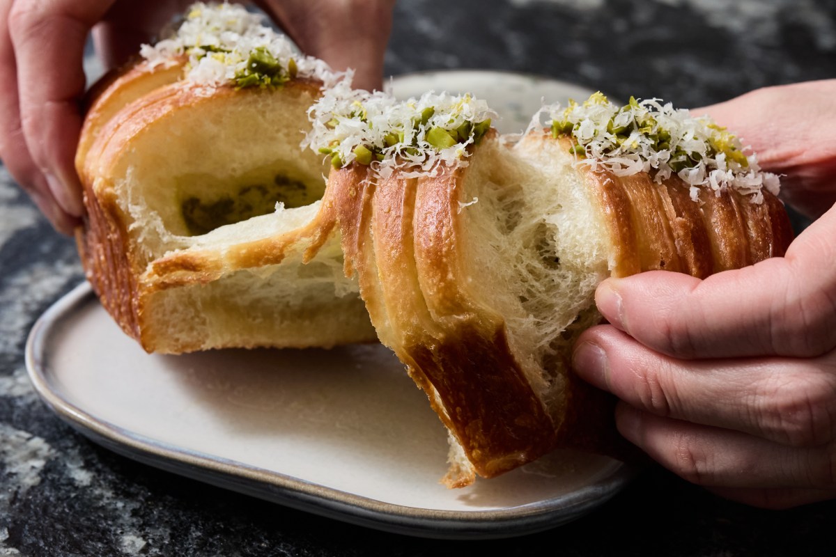 Close-up of hands pulling apart a golden brioche-style bun topped with pistachio and citrus zest on a plate.