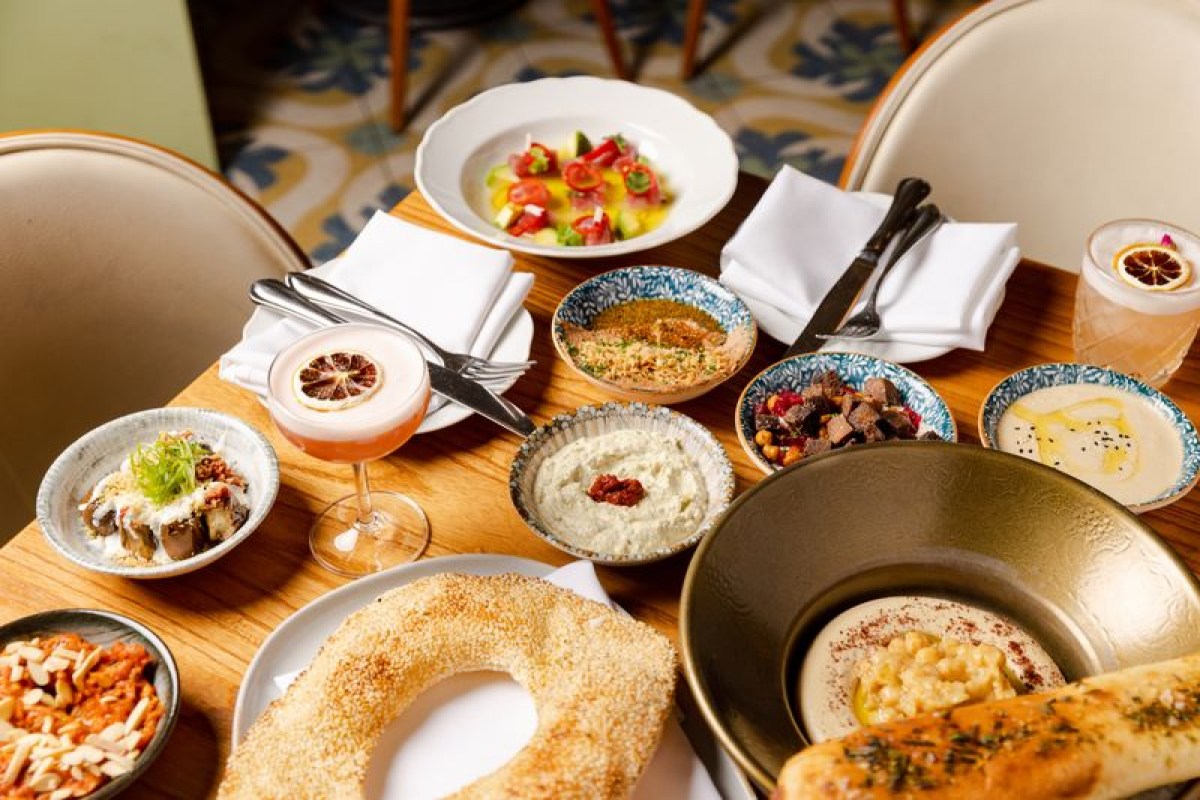 Overhead view of a wooden table set with multiple small plates of Mediterranean mezze, bread, and cocktails