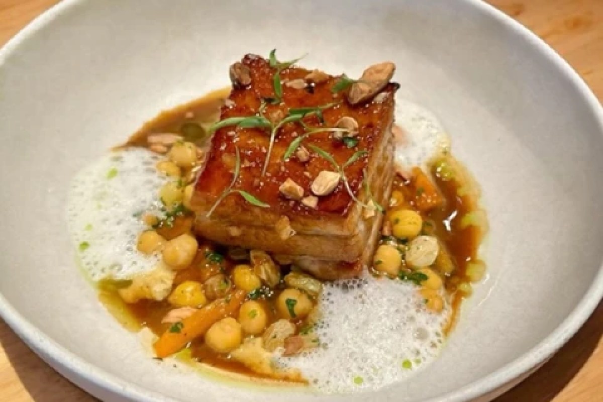 Plated dish of glazed pork belly with chickpeas, carrots, herbs and foam in a white bowl.