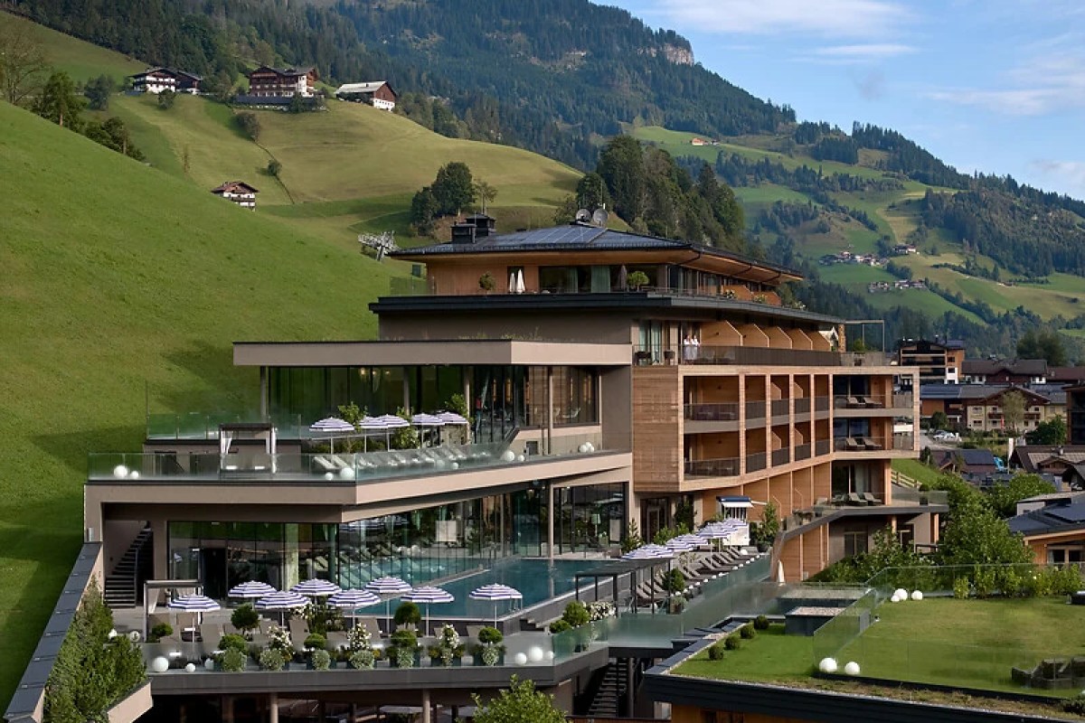 Modern wood-and-glass hotel building with terraces, pool and umbrellas set against green alpine hillside
