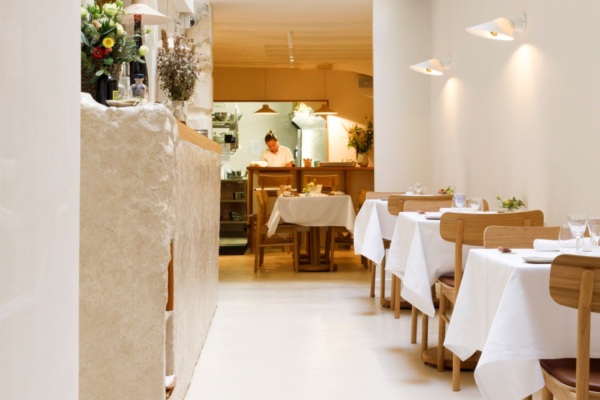 Minimalist restaurant dining room with white-clothed tables, wood chairs, stone wall, and chef working in open kitchen background
