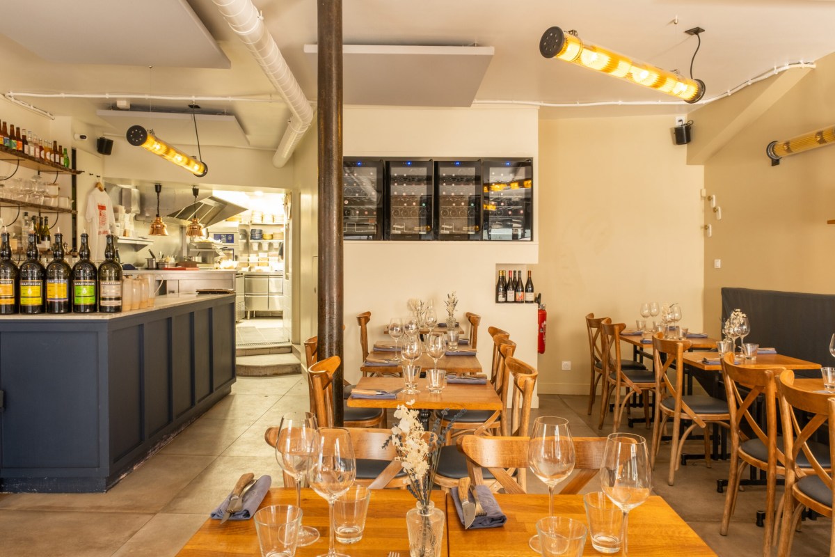 Restaurant dining room with wooden tables set for service, bar counter with wine fridge and open kitchen visible in background