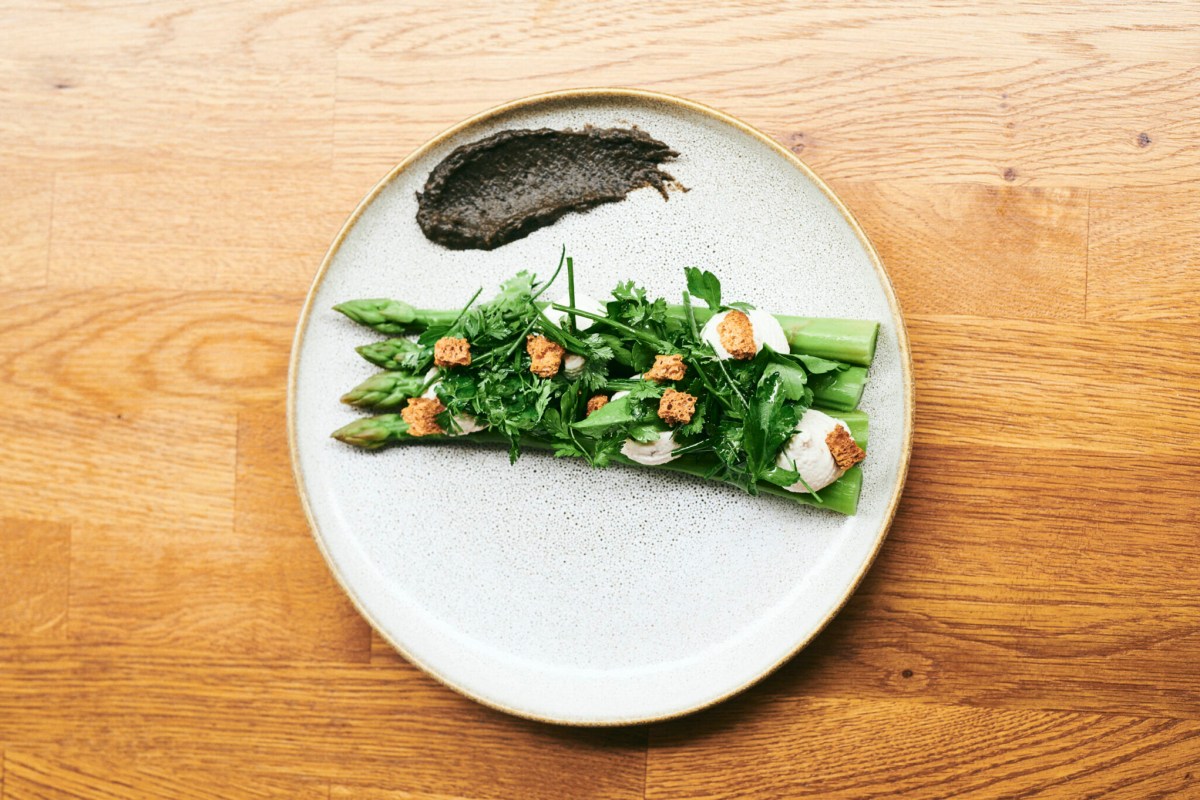 Plated dish of asparagus spears topped with herbs and croutons alongside a dark sauce swirl on a wooden table