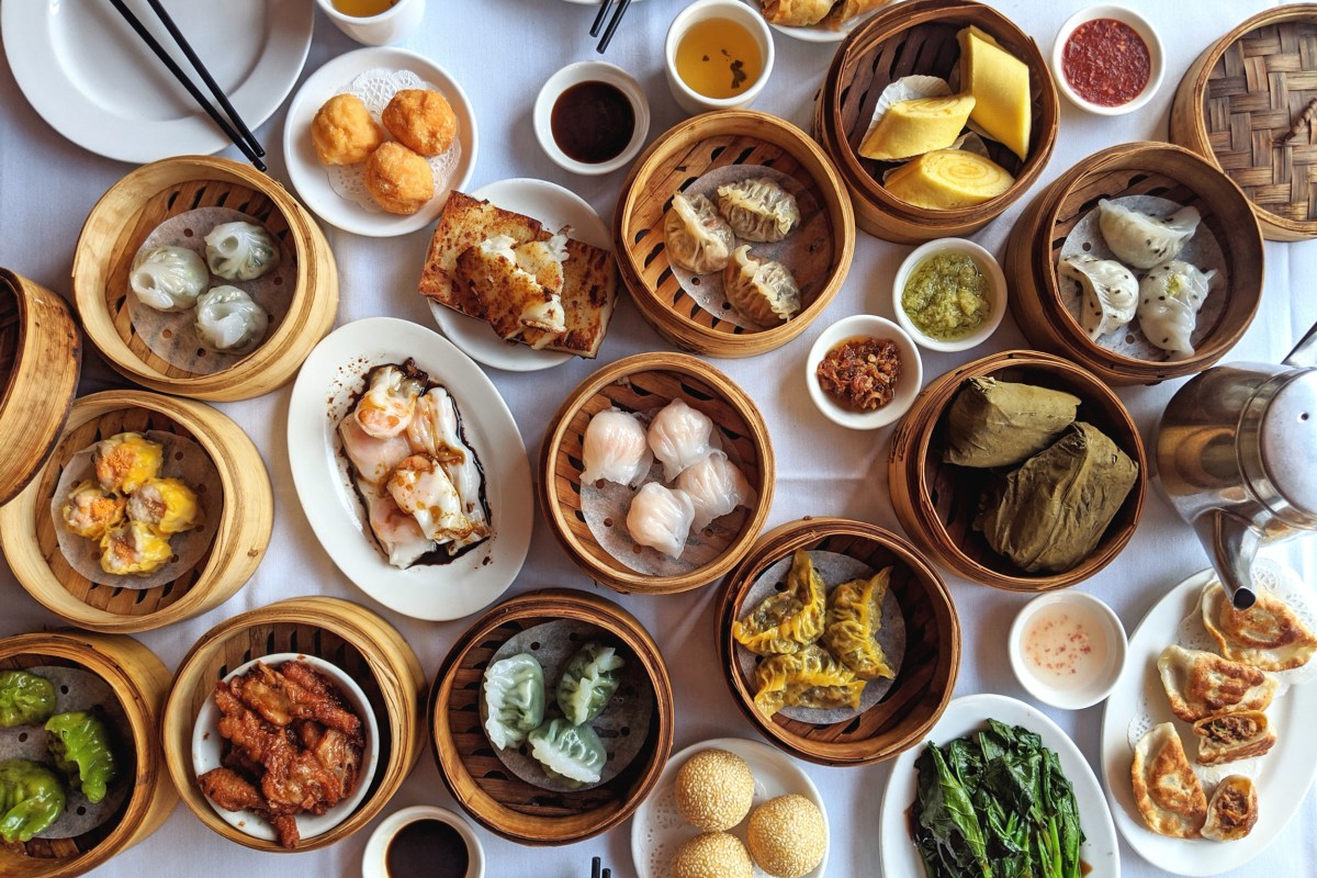 Overhead view of many bamboo steamer baskets and plates with assorted dim sum dishes on a white tablecloth