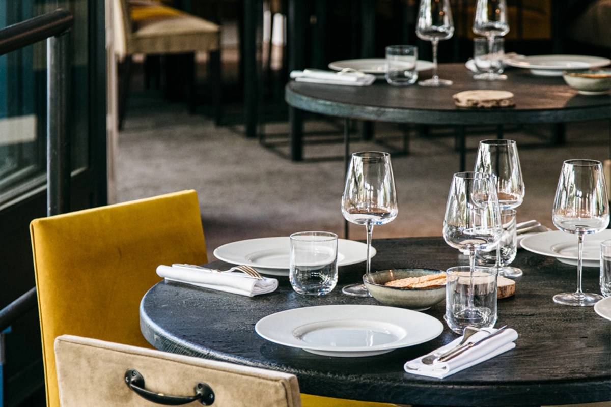 Round dark wood tables set with white plates, wine glasses, and yellow upholstered chairs in the dining room