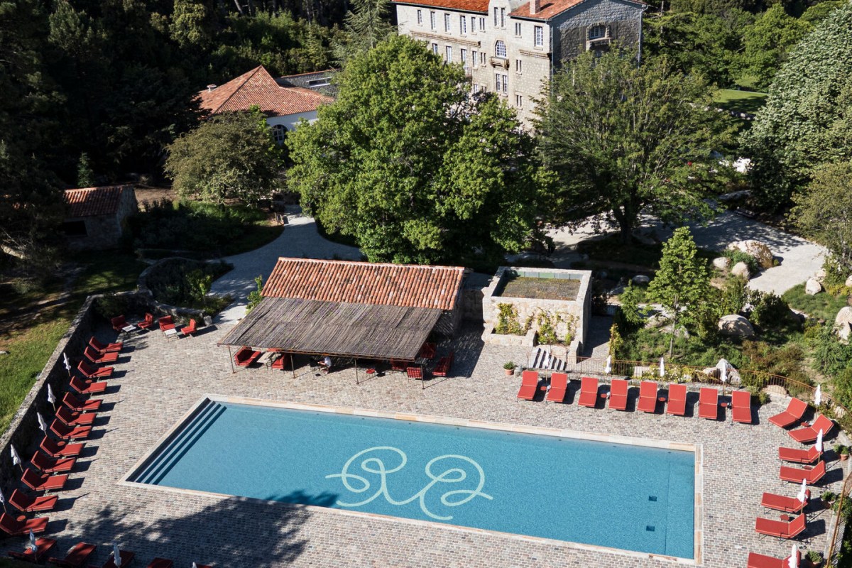 Aerial view of hotel façade, pool with monogram, red loungers and gardens