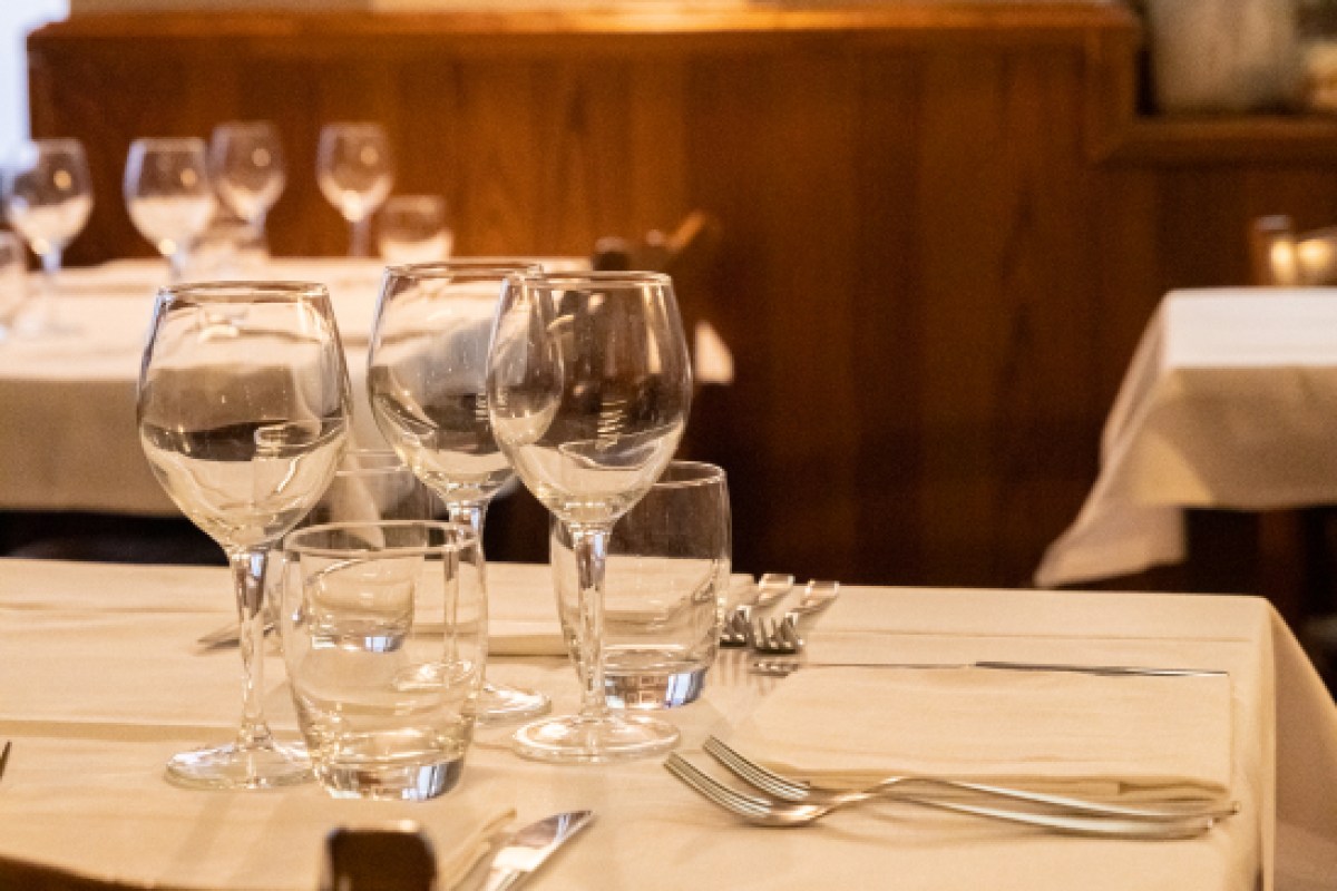Close-up of a set restaurant table with wine and water glasses, cutlery, and white tablecloth in a warm dining room.