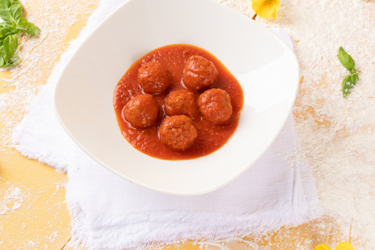 White bowl of meatballs in tomato sauce on a napkin with basil leaves and flour dusting.