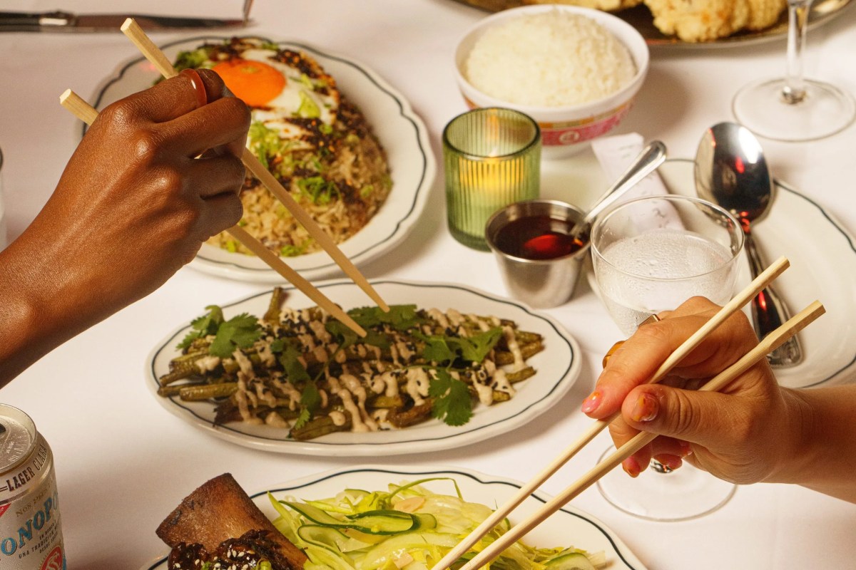 Close-up of hands using chopsticks over a table spread with fried rice, okra dish, and condiments