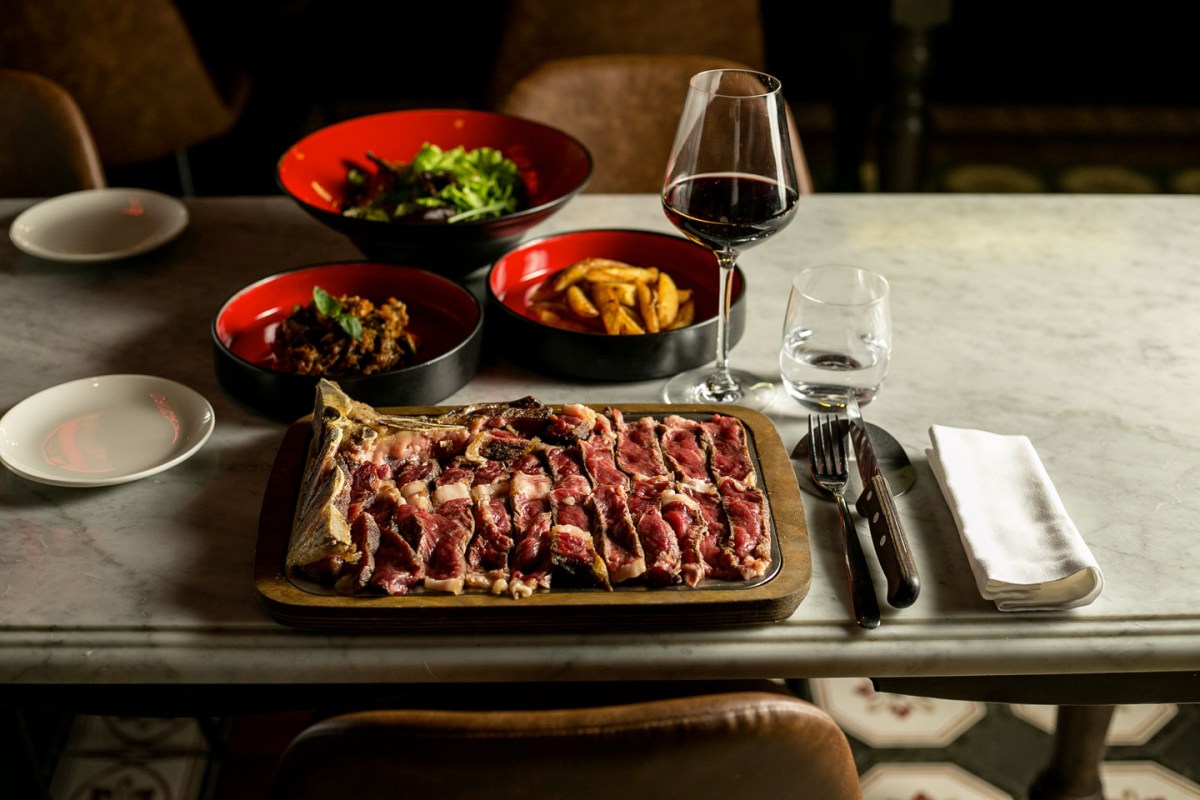 Table set with sliced dry-aged steak on wooden board, side dishes, red wine glass and cutlery