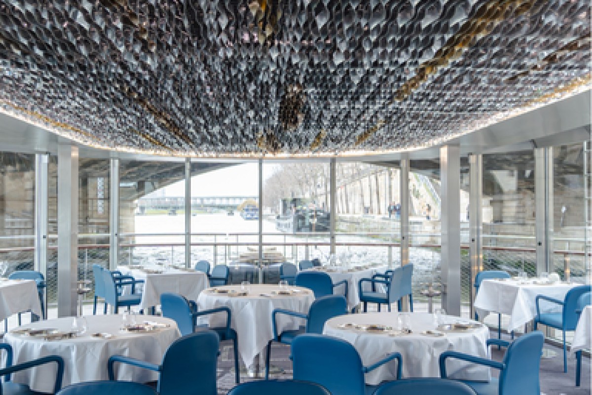Dining room aboard Ducasse sur Seine with blue chairs, set tables and a textured ceiling, river view through windows