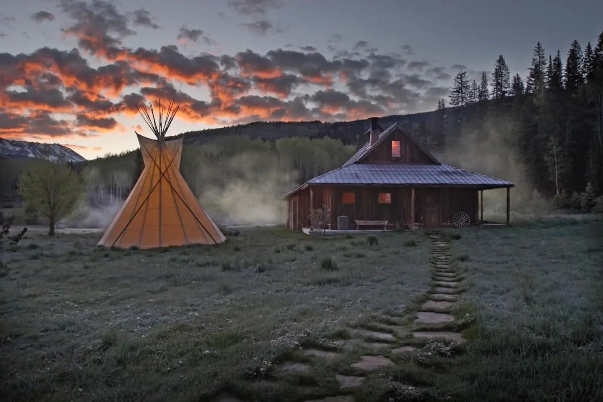 Rustic wooden cabin at dusk with glowing window, teepee beside it, stone path leading up through misty meadow under colorful sunset sky