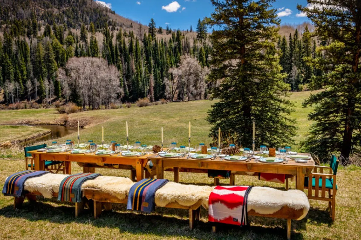 Long outdoor dining table set with plates and candles, surrounded by mountains and forest, at Dunton Hot Springs.