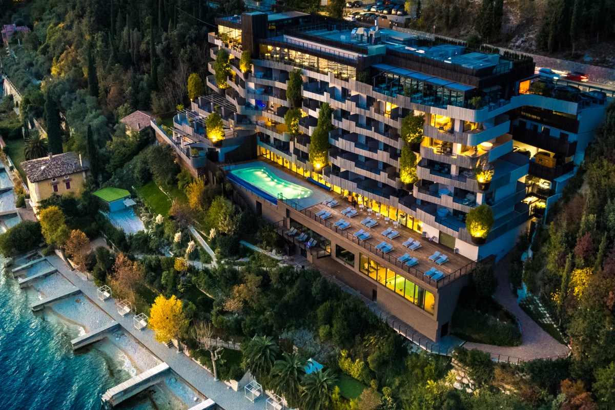 Aerial night view of the hotel building lit up with illuminated infinity pool, terrace, and Lake Garda shoreline below.