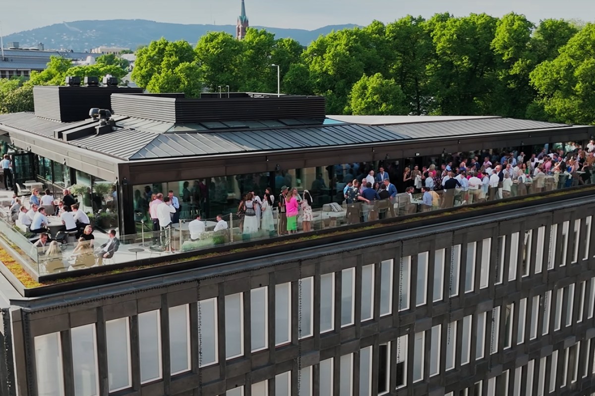 Aerial view of Eero's rooftop terrace with guests, glass railings, and Oslo skyline with trees and hills in background