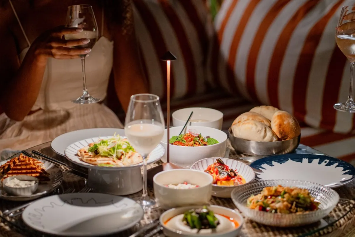 Table set with Lebanese mezze dishes, bread, and dips at an outdoor dining setting with wine glasses, striped cushions in background.