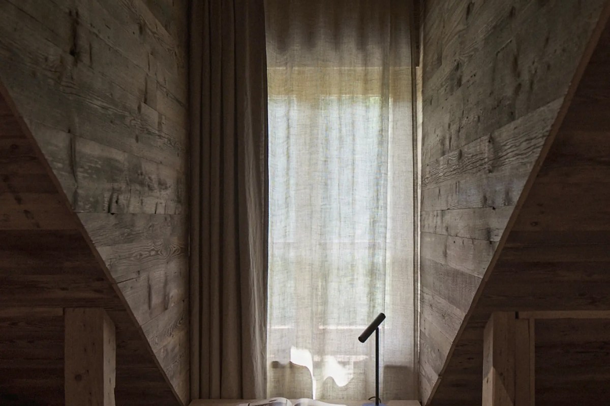 Minimalist wood-paneled guest room nook with sheer curtain, soft daylight, and a small reading lamp on a table.