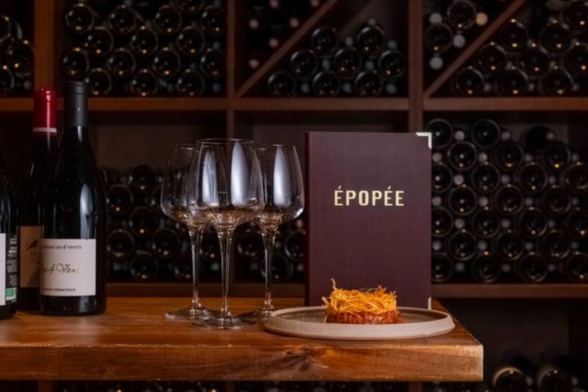 Wine cellar display with glasses, wine bottles, the restaurant's branded menu reading 'ÉPOPÉE', and a plated dish on a wooden counter