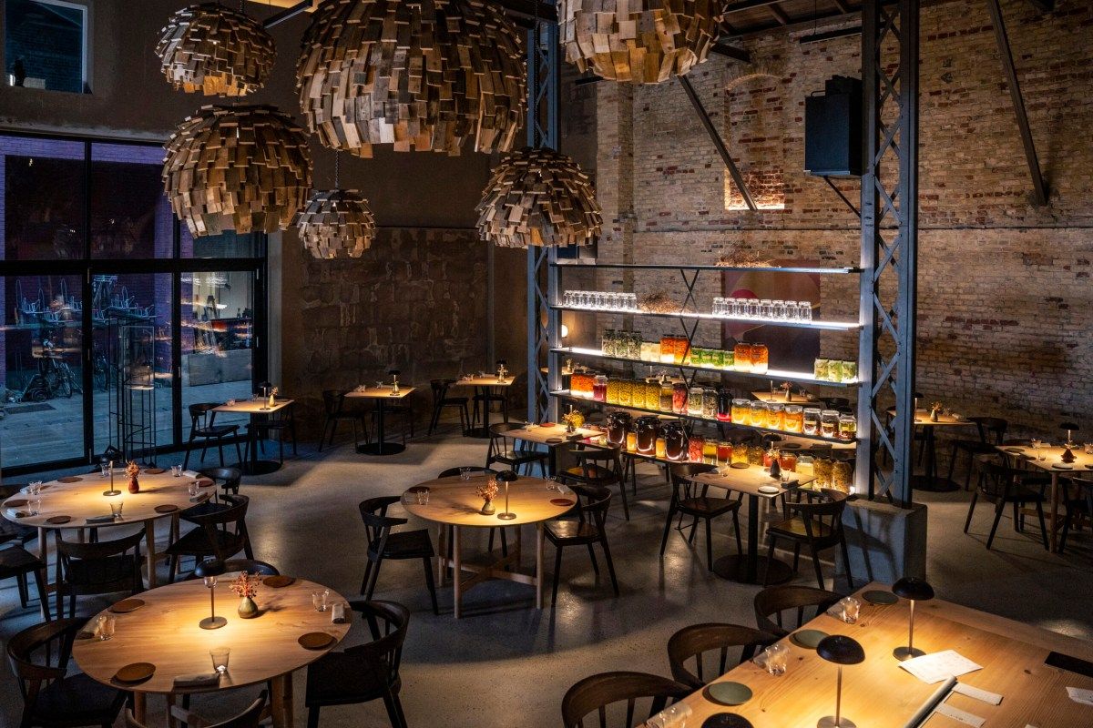 Full restaurant dining room with wood tables, dark chairs, sculptural pendant lights and a jar-filled shelf, view toward windows