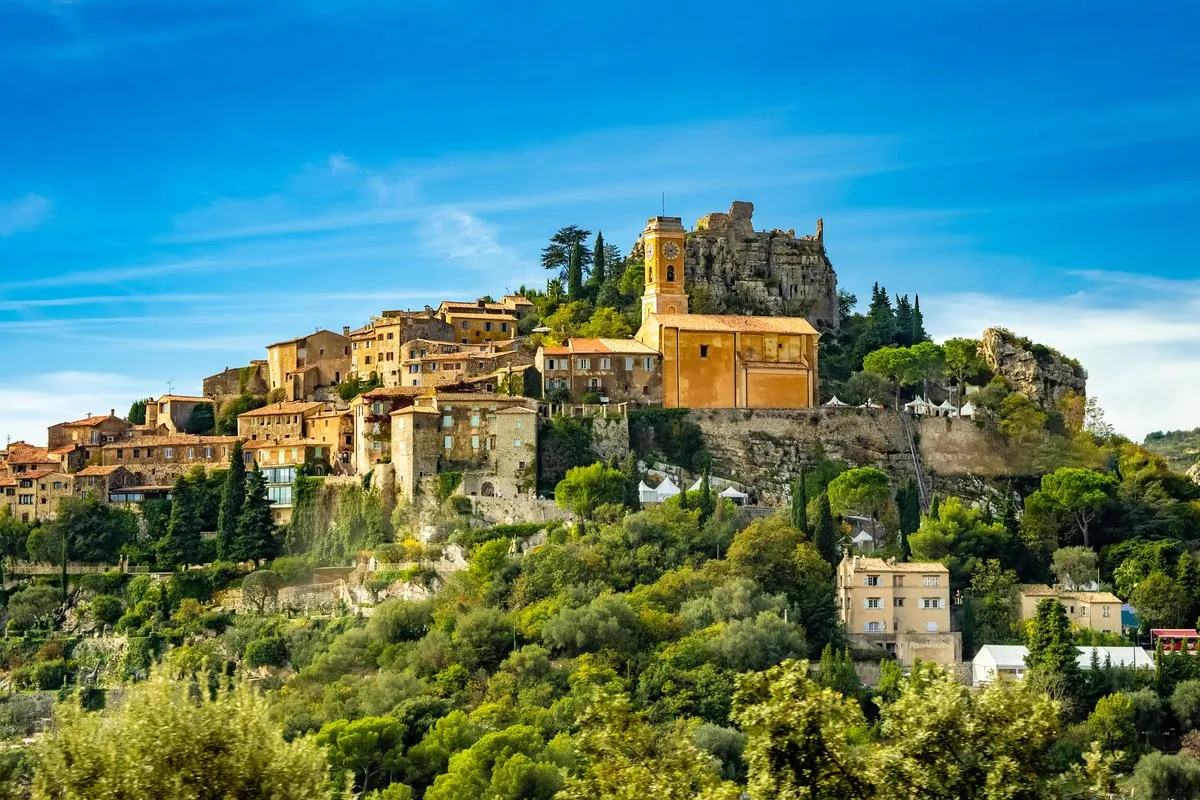 The medieval village of Èze with its ochre-yellow church tower on the rock, surrounded by greenery under a blue sky