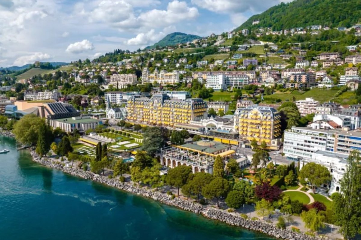 Aerial view of Fairmont Le Montreux Palace's yellow façade along Lake Geneva, surrounded by gardens and hillside town.