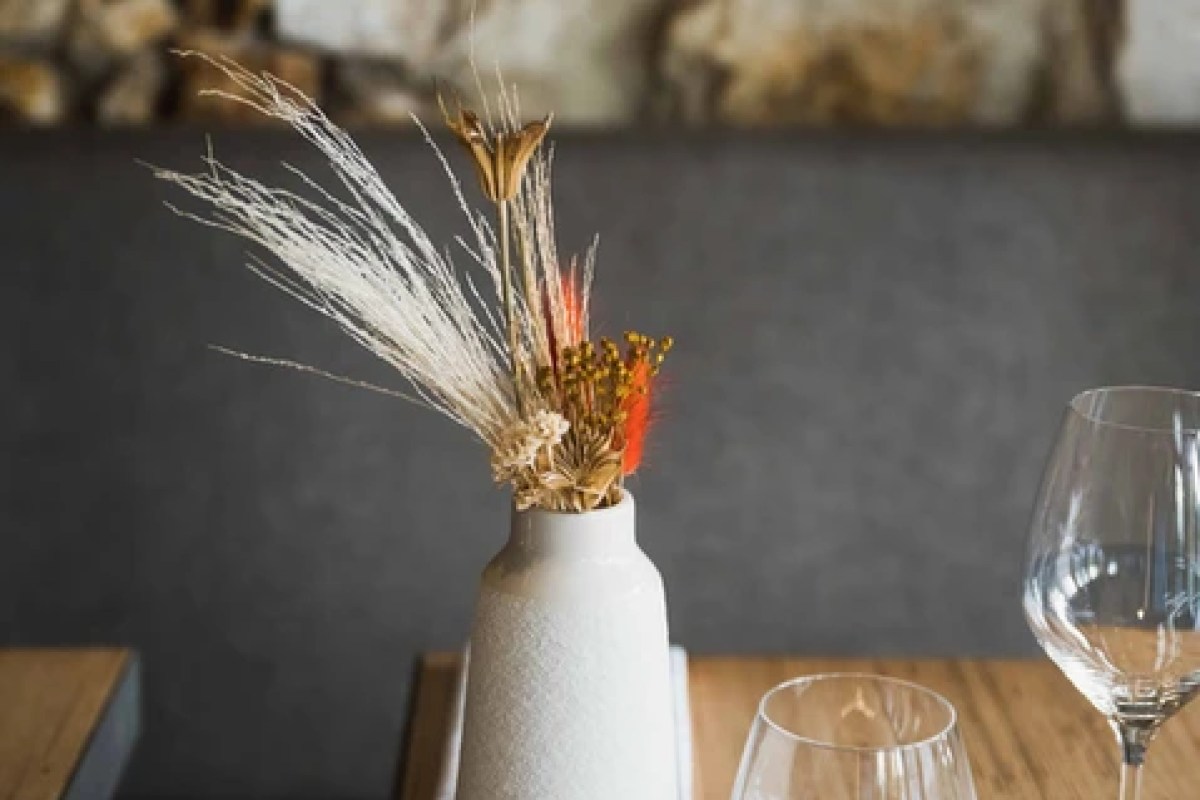 Dried flower arrangement in a white vase on a restaurant table with wine glasses and stone wall backdrop