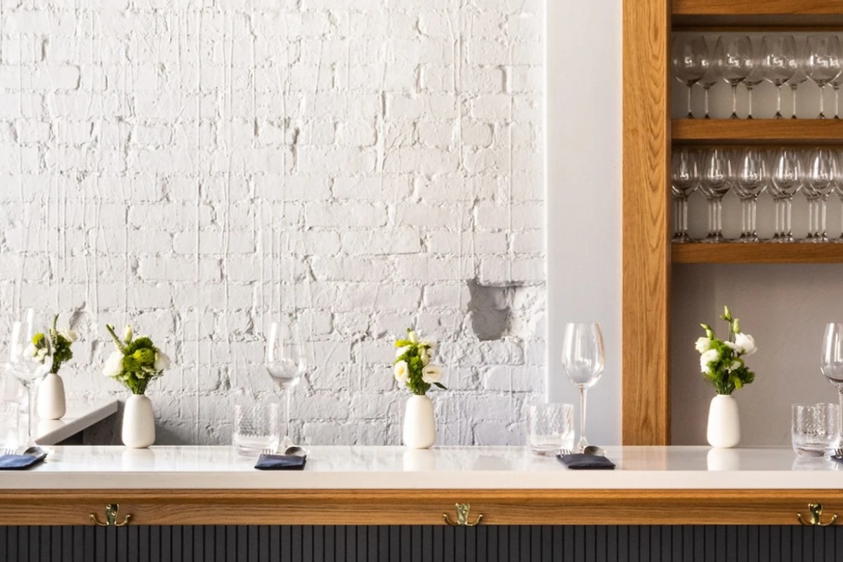 Same bar counter view with flowers, wine glasses, and shelving of stemware along a white brick wall