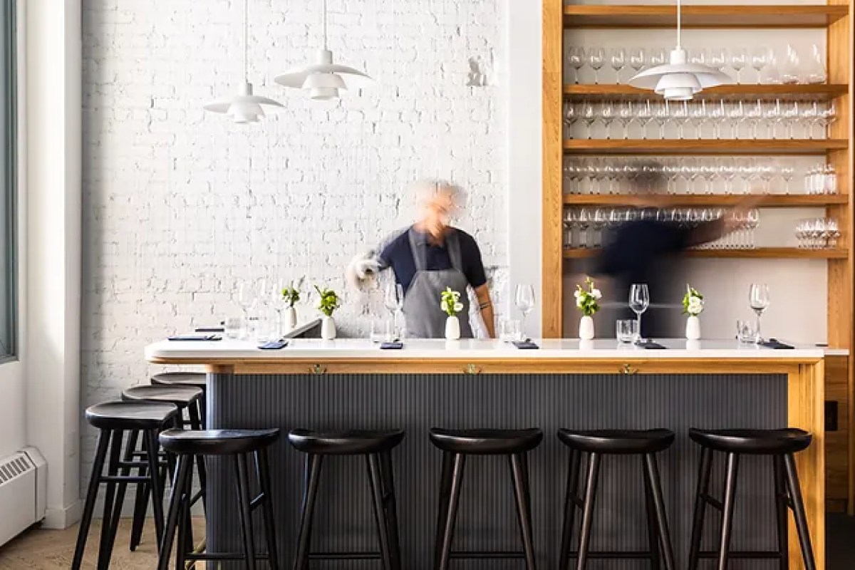 Wide view of the bar area with black stools, pendant lights, glassware shelves, and staff blurred in motion behind the counter