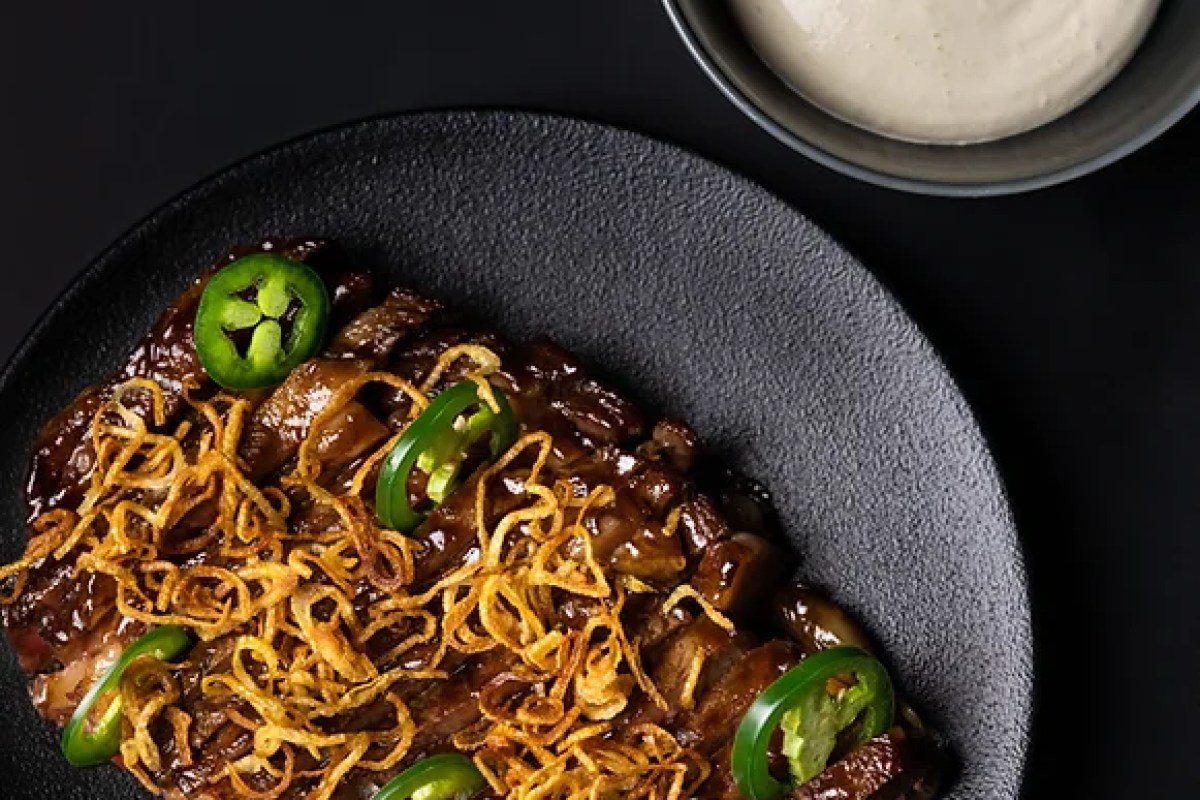 Dark glazed meat dish topped with crispy fried onions and jalapeño slices, served with a side sauce