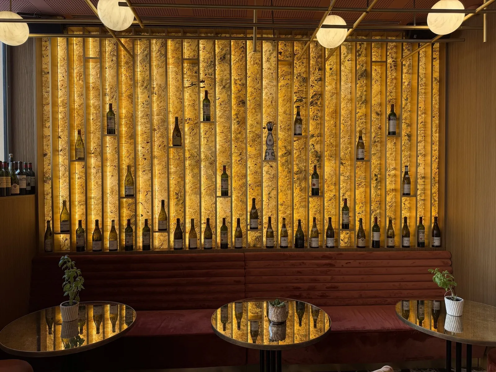 A backlit amber wall of wine bottles on brass ledges above a terracotta velvet banquette at Ferni in Antibes