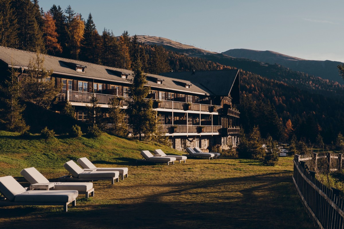 Modern wooden hotel building with balconies on a mountainside, sun loungers on lawn, forested peaks behind