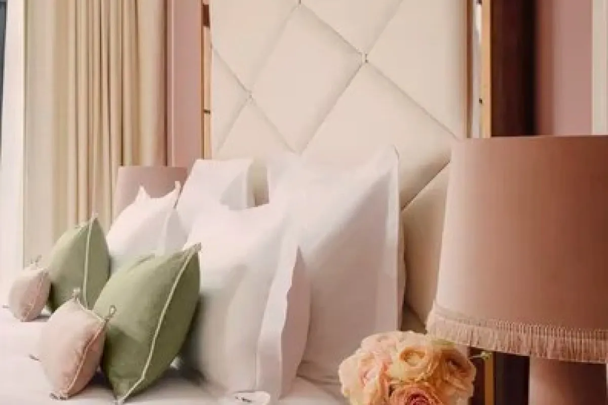 Close-up of a quilted cream headboard with pink and green pillows and a blush lamp, suggesting a hotel guest room.