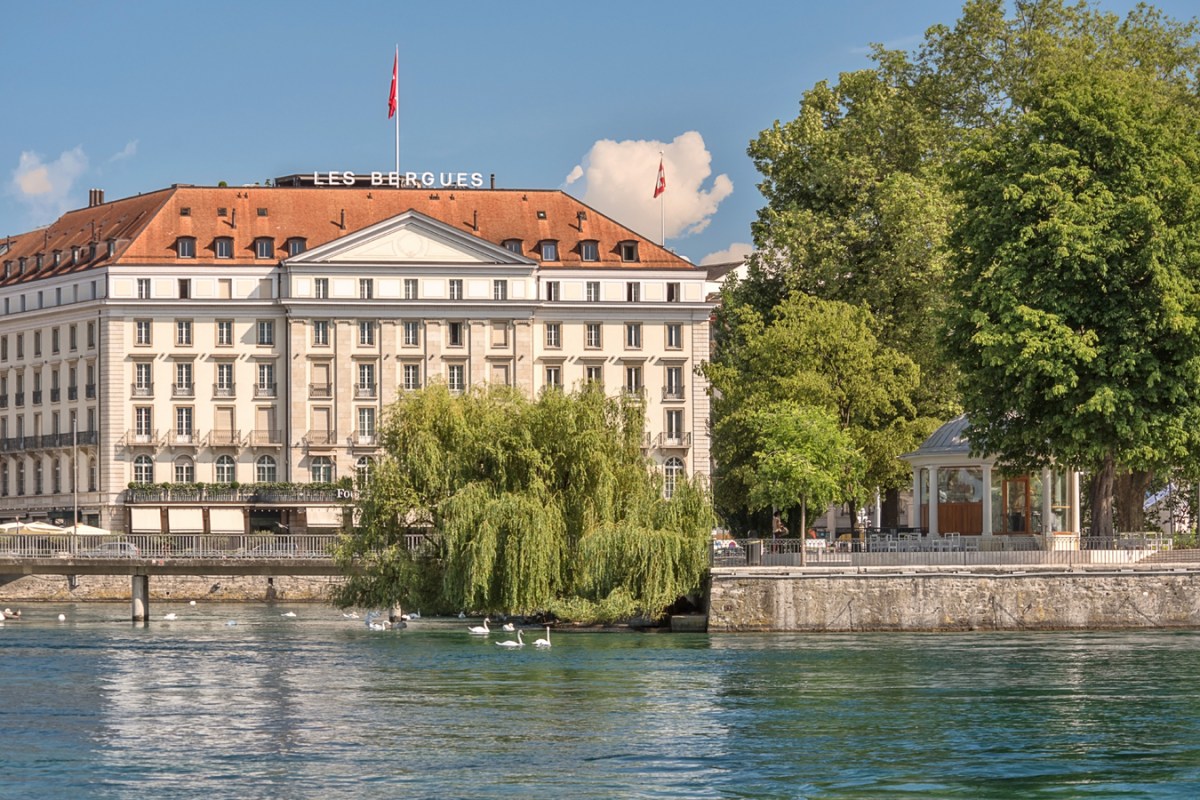 Exterior façade of Four Seasons Hotel des Bergues Geneva seen across the river with swans, trees and clear sky