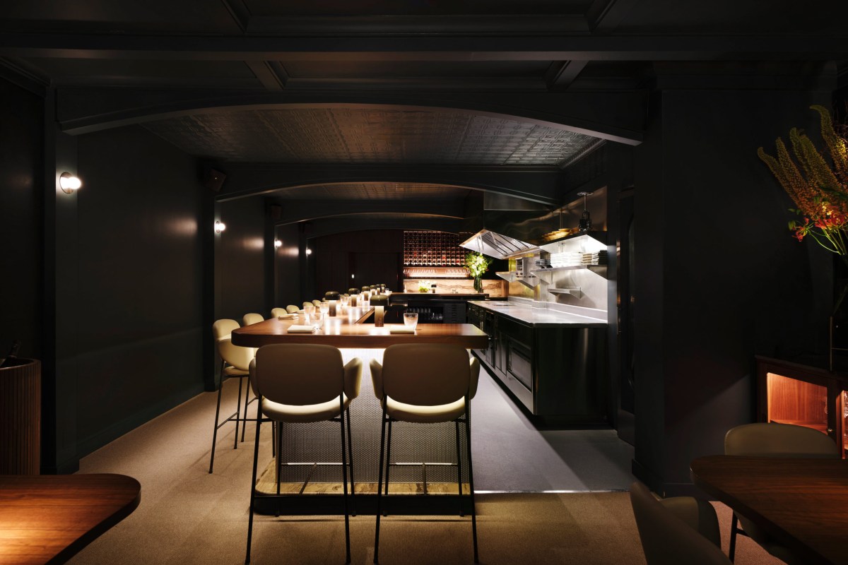Dark, elegant dining room with a long wooden bar, cream stools, and an open kitchen in the background