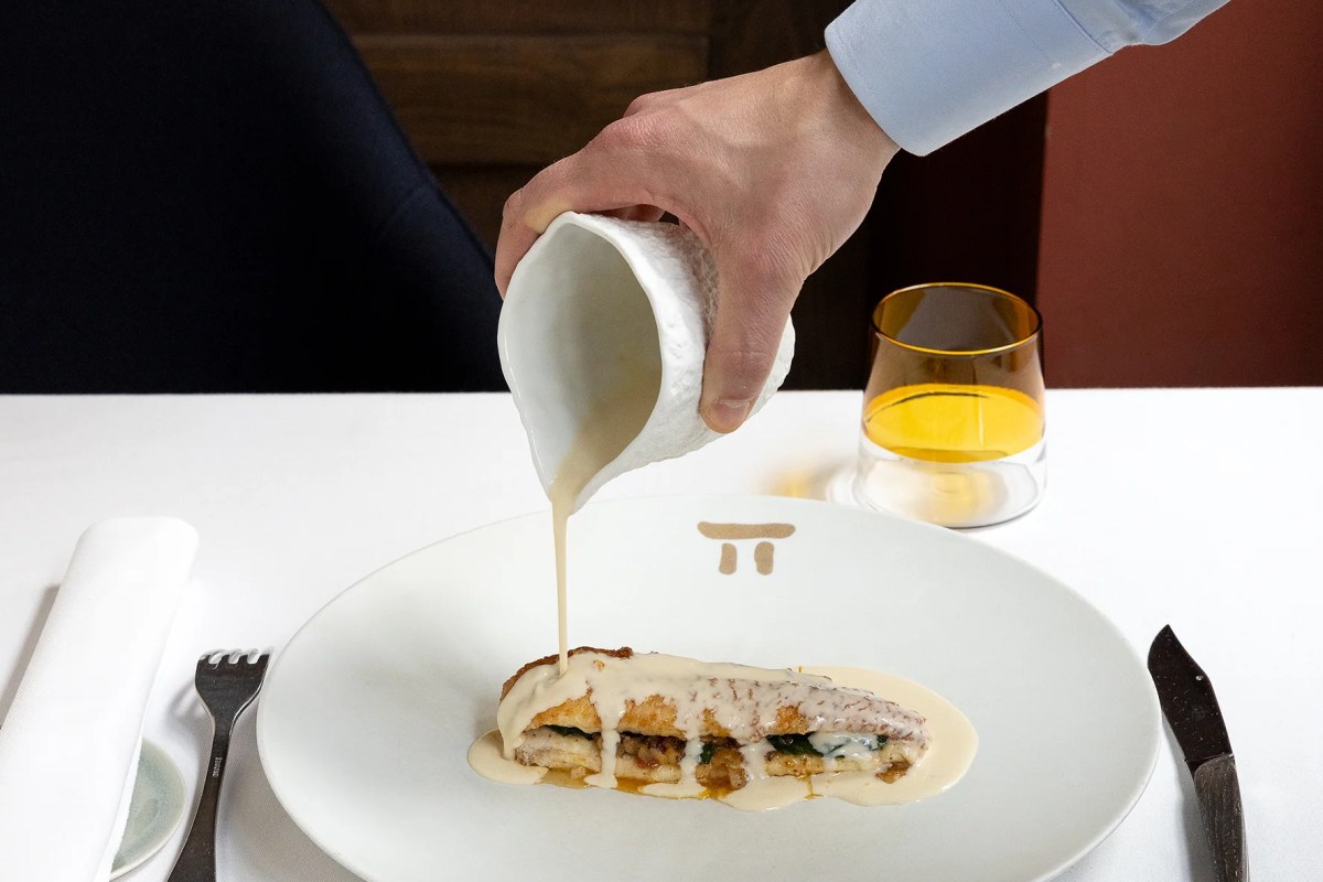 Waiter's hand pouring a creamy sauce over a plated fish dish at the table, with a glass of wine beside it