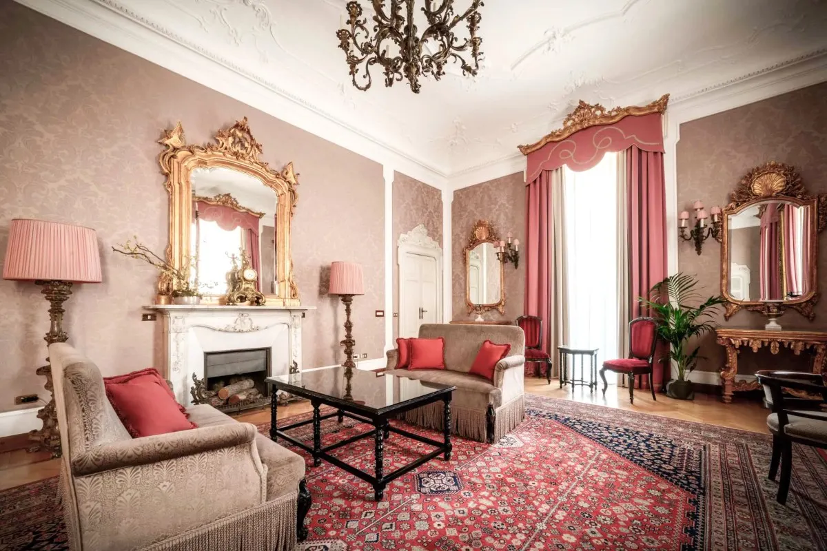 Opulent hotel suite living room with gilded mirrors, fireplace, and red patterned rug