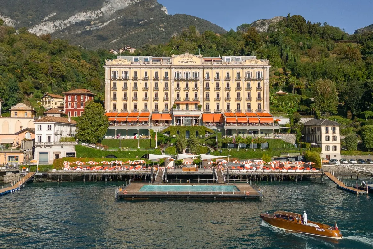Daytime view of Grand Hotel Tremezzo's cream façade from the lake, with floating pool, boat, and mountains behind.