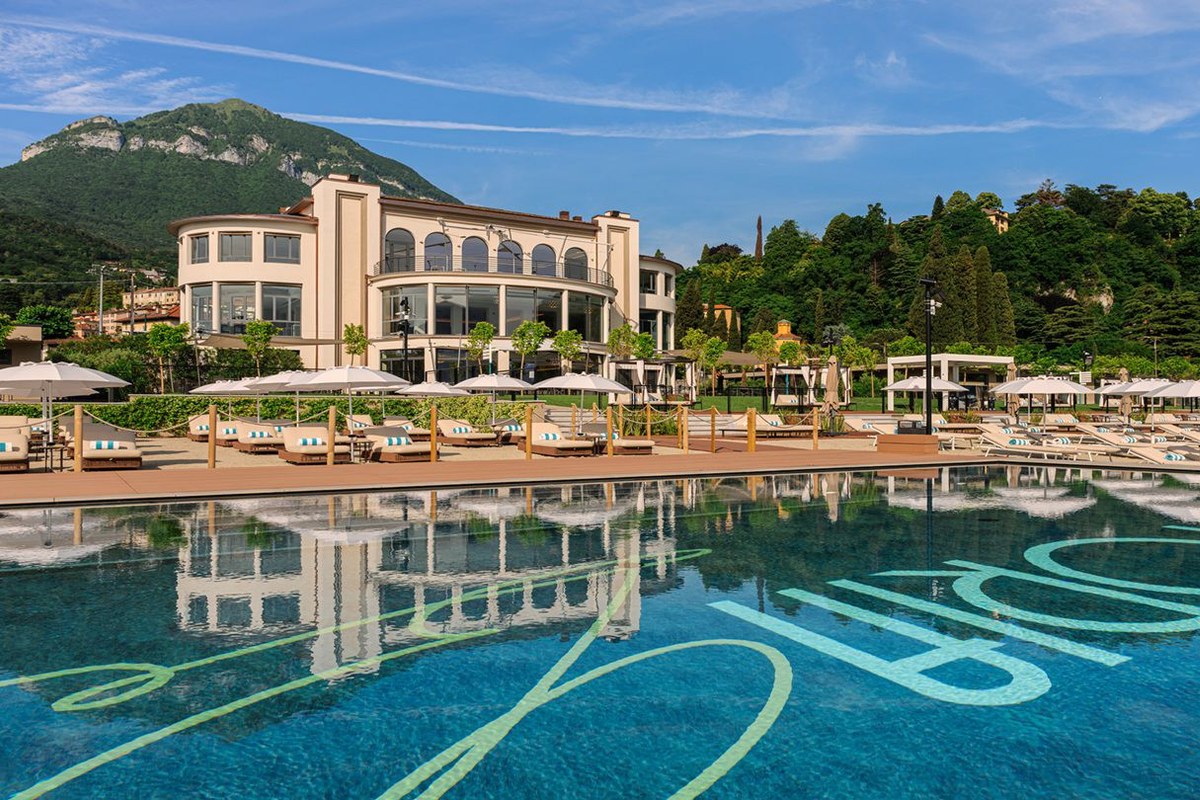 Infinity pool with loungers and umbrellas in front of hotel building and mountain backdrop