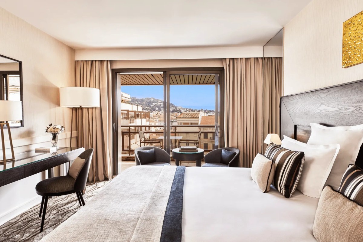 Hotel guest room with bed, desk, and balcony offering a view of hills and distant sea